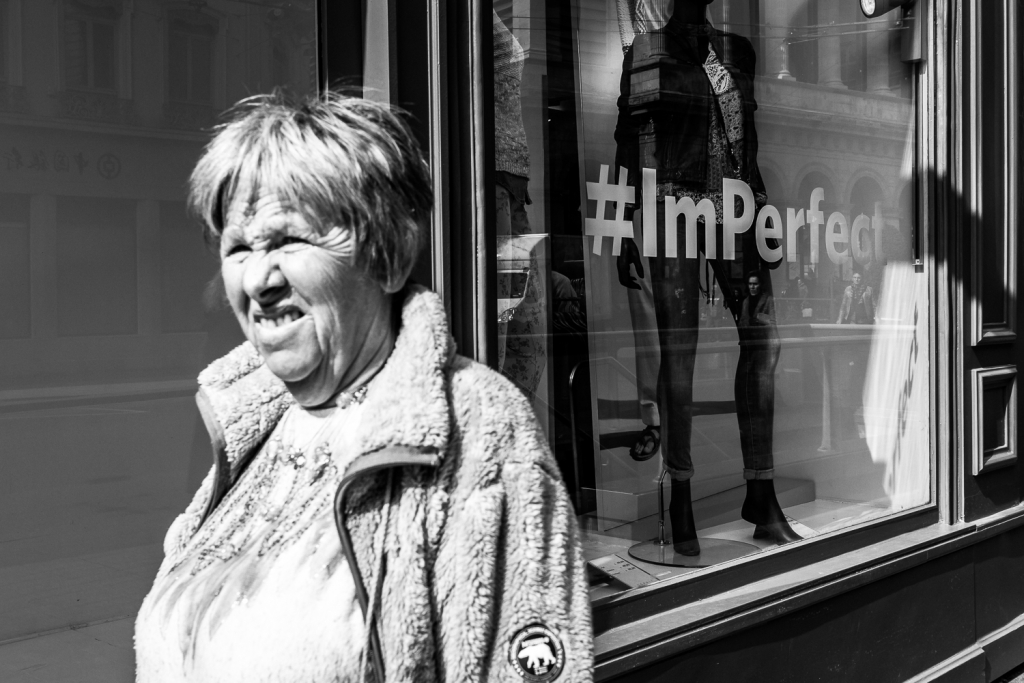Black and white photo. A woman with a wrinkled face in her later years, modestly dressed. Her appearance leads us to believe that she may be living on the street. In the background we see a shop window with skinny models and a hashtag saying I am prefect