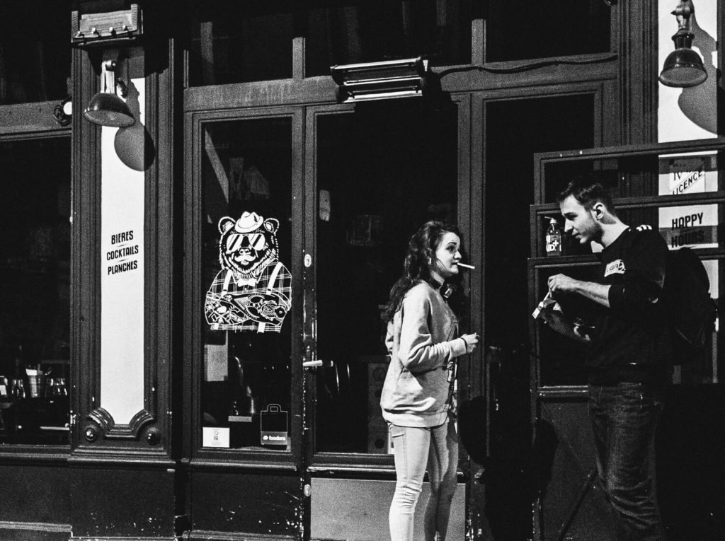 Black and white photo. A man and a woman are talking in front of a bar. A woman holds a cigarette in her mouth while a man is taking a cigarette out of a pack. The man tenderly observes the woman and smiles.