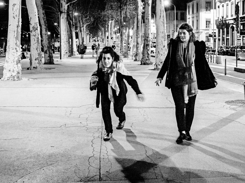 Black and white photo. Two girls on the streets of Lyon. The girl on the left holds a plastic cup with a drink in her hand and stumbles