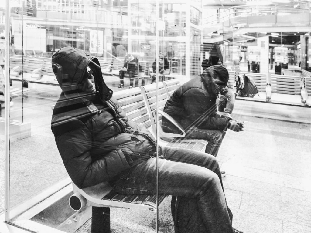 Black and white photo. Behind the Plexiglas, in the foreground we see a male person sleeping leaning against the glass. His head and face are hidden with a cap and hood. Behind him in the background we can see a number of other people who are also sleeping.