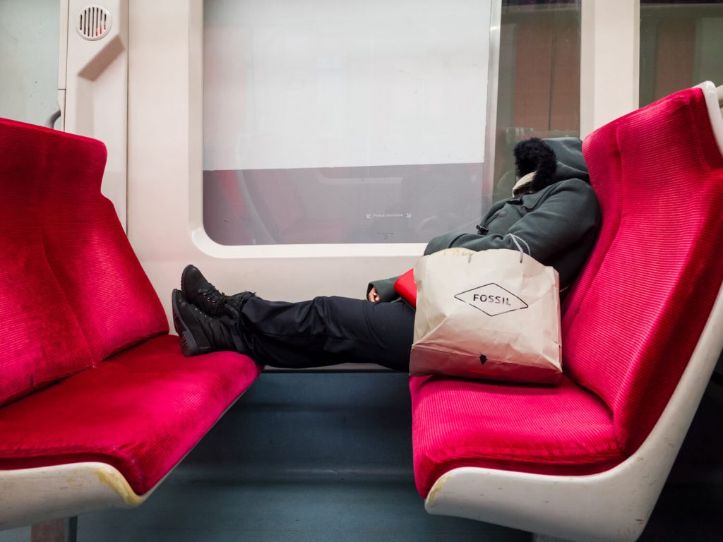 Red metro seats facing each other. At the bottom of the seat, a person in a thick winter jacket with a hood on his head is sleeping. Her body is stretched across both seats. The legs are on one seat while the body is on the other. The impression is that the person is sleeping. 