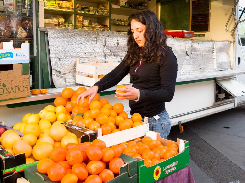 A girl in a black jacket arranges oranges at the market.  Her long curly hair highlights her face illuminated by the warm morning sun