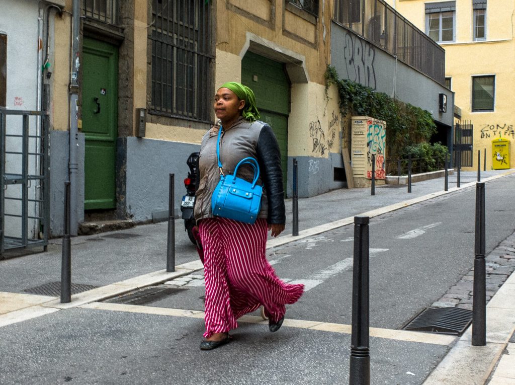 A woman is walking down the street.  She wears a long red skirt with lines. The skirt flutters from the energetic movement. Her look is completed with a gray black jacket, a blue bag and a green scarf on her head.