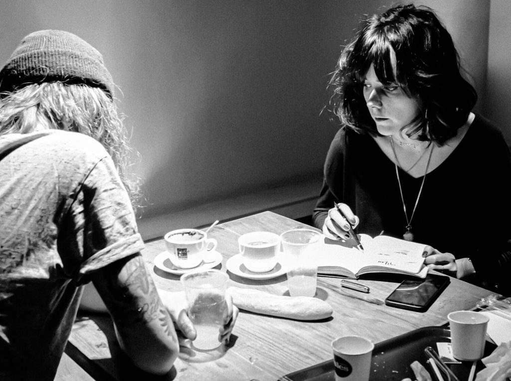 Black and white photo. A couple is sitting at a table in a cafe. On the table are empty coffee cups, juice cups, and part of a baguette. A girl holds a ballpoint pen above an open notebook while looking away