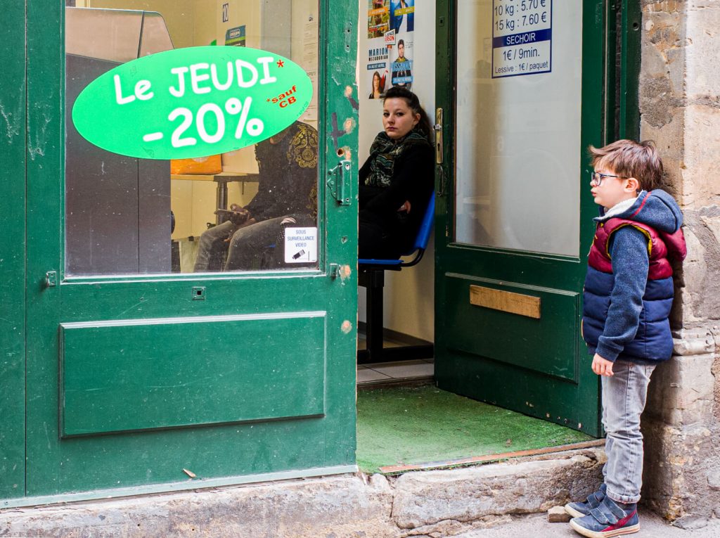 Automatic laundry shop. Through the open green door we see a woman sitting, waiting and looking directly into the camera lens.  In front of the door, a boy peers into the interior of the laundry room. There is a large sticker on the door with the inscription: "On Thursday 20% discount"