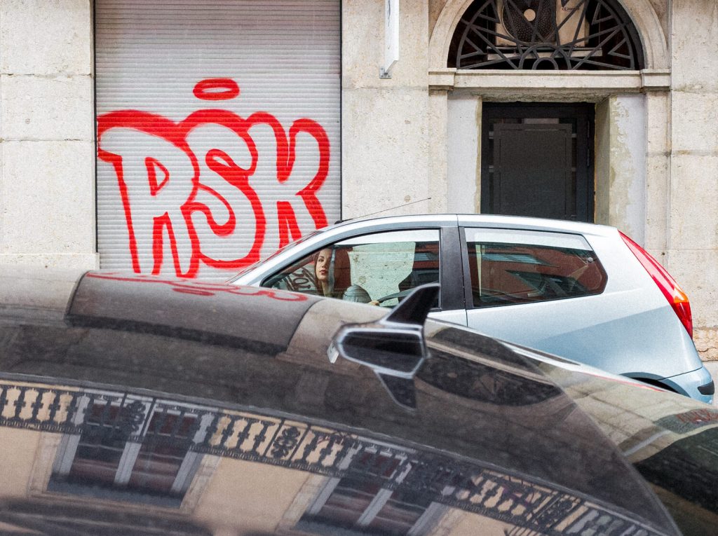 Three-layer photo. In the foreground is the hood of a black car that reflects the white windows on the orange facade. In the second plan is a girl in a gray car who starts the car and looks at the camera lense. In the background, there is a large red graffiti on the building wall that says RSK