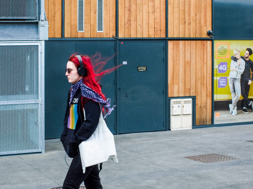 A girl in a black hoodie with bright red hair and headphones on her ears is walking down the street. The wind lifts her hair and highlights the red color of her hair against the blue door in the background