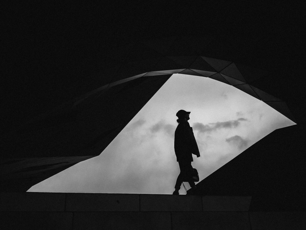 Black and white photo. The building frames the photo. In the center is a large opening through which we see the sky, clouds in the background, and the silhouette of a female person clutching a bag while looking at the sky