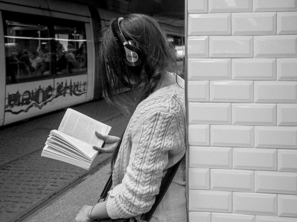 Black and white photo. A girl with headphones on her head and a book in her hand is leaning on a pillar covered with white tiles, waiting for the tram.