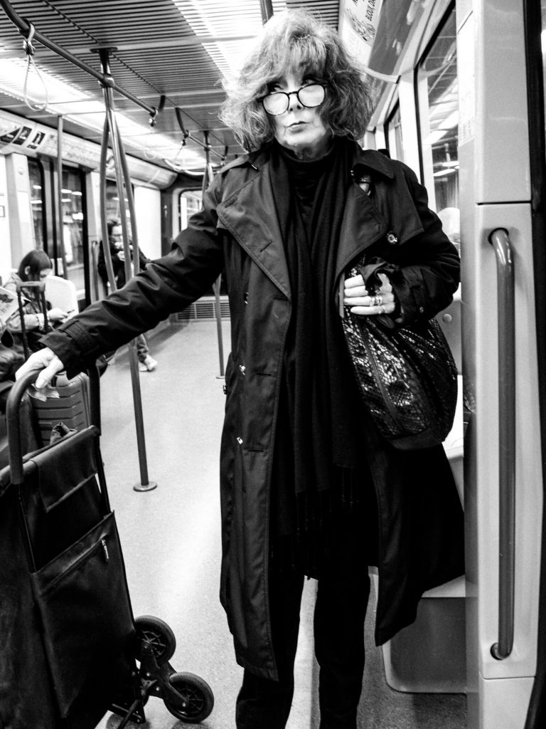Black and white photo. The interior of the subway train. A woman all in black, with bouffant hair. She clutches the bag with one hand while holding the handle of a large bag on wheels with the other. Her expression is stern as she looks over her glasses to the right