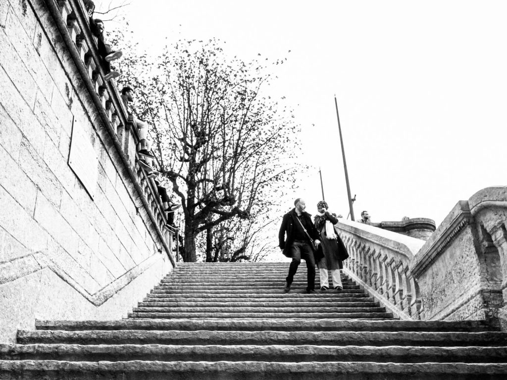 Black and white photo taken at the bottom of the stairs. The stairs direct the view upwards. There is a couple at the top. The man is still in motion as he looks worriedly in the direction of the woman. The woman has stopped and is holding onto her eye 