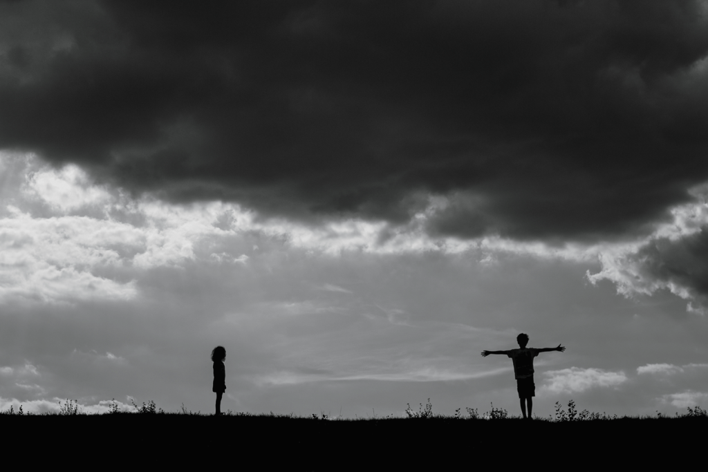 Black and white photo. Silhouettes of a boy and a girl standing on a hill. They are several meters away from each other. Heavy dark clouds descend above their heads. The boy is spreading his arms towards the sky, while the girl is standing facing the boy in a frozen pose.